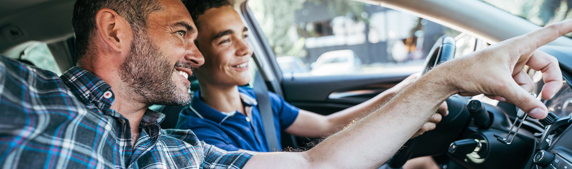 Ein lächelnder Mann (Beifahrer) und ein lächelnder junger Mann (Fahrer) sitzen in einem Auto, der Mann zeigt mit dem Finger auf etwas, das der junge Mann beachten soll